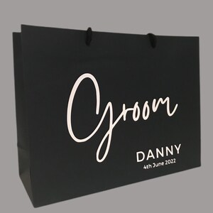 May include: Black gift bag with the word "Groom" in white script. Below the word "Groom" is the name "Danny" and the date "4th June 2022" in a simple, white font. The bag has black rope handles.