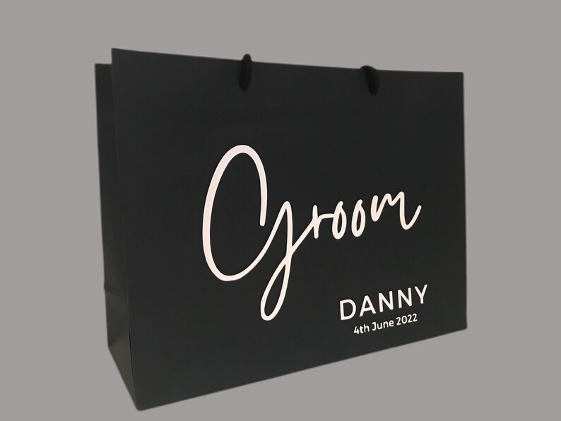 May include: Black gift bag with the word "Groom" in white script. Below the word "Groom" is the name "Danny" and the date "4th June 2022" in a simple, white font. The bag has black rope handles.