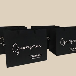 May include: Black gift bags with white text that reads "Groomsman" in a script font. One bag has the name "Ciaran" and the date "4th June 2022". Another bag has the name "Richard" and the date "4th June 2022".