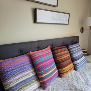 Peut inclure: Quatre coussins de canapé en crochet colorés avec des motifs rayés dans des tons de rose, violet, bleu, vert, jaune, orange et marron. Les coussins sont disposés sur un couvre-lit blanc à motif texturé.