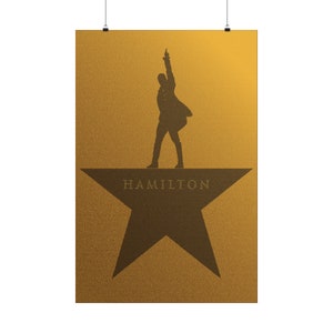 May include: A gold poster with a silhouette of a person standing on a large brown star. The star has the word "HAMILTON" written on it.