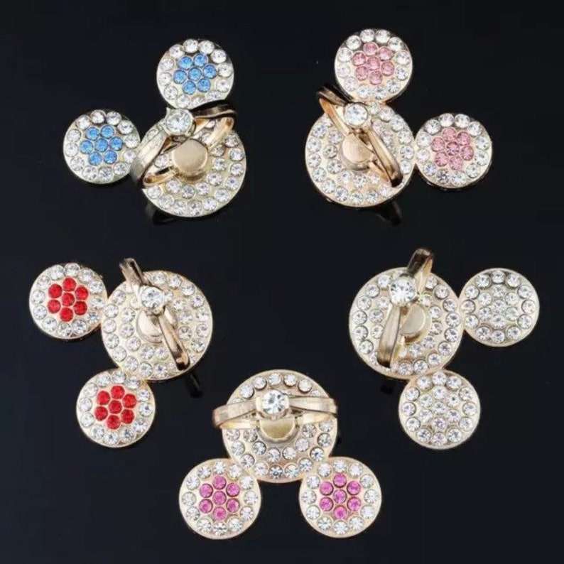 Może przedstawiać: Zestaw czterech uchwyt&oacute;w na pierścienie do telefonu z wzorem Myszki Miki. Każdy uchwyt ma inny kolor środka z kryształk&oacute;w i przezroczysty pierścień.