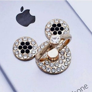 Może przedstawiać: Złoty uchwyt do telefonu z obrotowym pierścieniem i trzema okrągłymi, błyszczącymi, czarno-białymi akcentami ozdobionymi kryształkami.