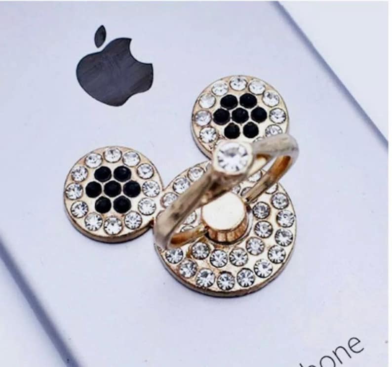 Może przedstawiać: Złoty uchwyt do telefonu z obrotowym pierścieniem i trzema okrągłymi, błyszczącymi, czarno-białymi akcentami ozdobionymi kryształkami.