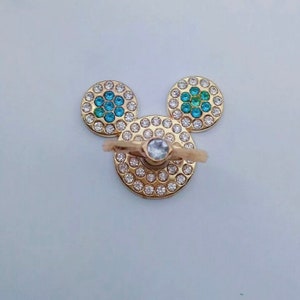 May include: Gold phone ring holder with a clear crystal in the center and two round, blue and green rhinestone accents on the sides, shaped like Mickey Mouse ears.