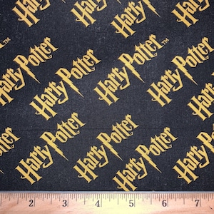 May include: Black fabric with a repeating pattern of the words "Harry Potter" in gold lettering. The fabric is measured with a ruler.