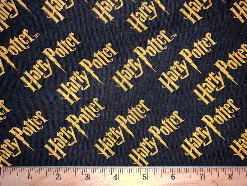 May include: Black fabric with a repeating pattern of the words "Harry Potter" in gold lettering. The fabric is measured with a ruler.