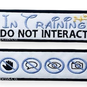 May include: Two rectangular white patches with black borders. The top patch reads "In Training DO NOT INTERACT" in blue and black embroidery. The bottom patch features four blue-outlined symbols: a hand, a speech bubble, an eye, and a camera, each crossed out.
