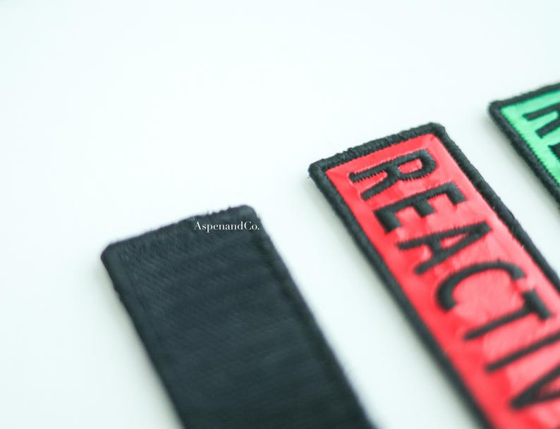 Three dog patches with black stitching. The first patch is showing the back hook side of the patch. The second patch is red with the text 'REACTIVE.' The third patch is green with the text 'REACTIVE.'