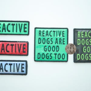 Group of five holographic dog patches displayed on a white background. Three rectangular REACTIVE patches in red, green, and blue are shown alongside two square REACTIVE DOGS ARE GOOD DOGS TOO patches in green holographic and black with green letters holographic. All patches have hook backing.