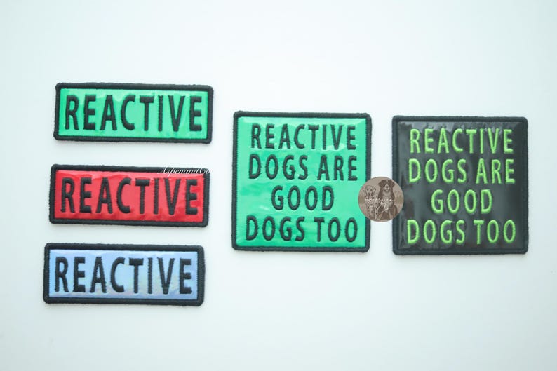 Group of five holographic dog patches displayed on a white background. Three rectangular REACTIVE patches in red, green, and blue are shown alongside two square REACTIVE DOGS ARE GOOD DOGS TOO patches in green holographic and black with green letters holographic. All patches have hook backing.