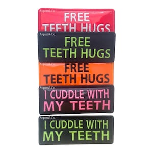 May include: A stack of five rectangular patches with humorous text. The patches are red, black, orange, pink, and black. The text on the patches reads "FREE TEETH HUGS" and "I CUDDLE WITH MY TEETH" in white and green.
