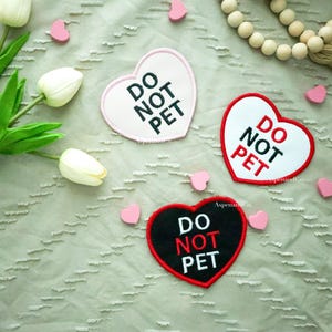 May include: Three heart-shaped patches with the text "DO NOT PET" in black and red. One patch is pink with a white border, another is white with a red border, and the third is black with a red border. Pink heart-shaped decorations and white tulips are scattered around the patches.