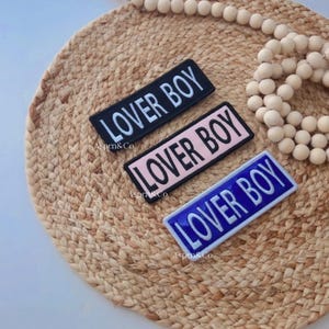 May include: Three rectangular patches with the text "LOVER BOY" in white. The patches are black, pink, and blue. The patches are on a woven, circular mat with a beaded garland.