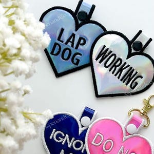May include: Heart-shaped pet tags with text: "LAP DOG", "WORKING", "IGNORE ME", and "DO NOT PET". The tags are iridescent, blue, and pink with white stitching and black borders. Each tag has a loop for attachment.