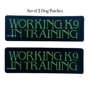 May include: Two black rectangular dog patches with green embroidered text. The text reads "WORKING K9 IN TRAINING". The patches also include the text "AspenandCo." at the bottom left corner. The image is labeled "Set of 2 Dog Patches".
