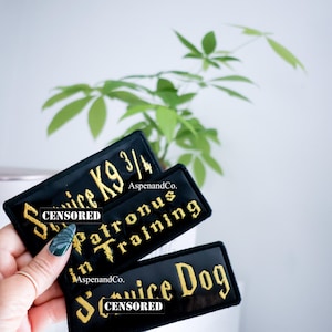 May include: Three black embroidered patches with gold lettering. The top patch reads "Service K9 3/4", the middle patch reads "in Patronus Training", and the bottom patch reads "Service Dog". The patches are from AspenandCo.