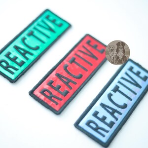 Three holographic REACTIVE dog patches in a 4 x 1.5 inch rectangle shape, shown in red, green, and blue colors. Each patch has hook backing.