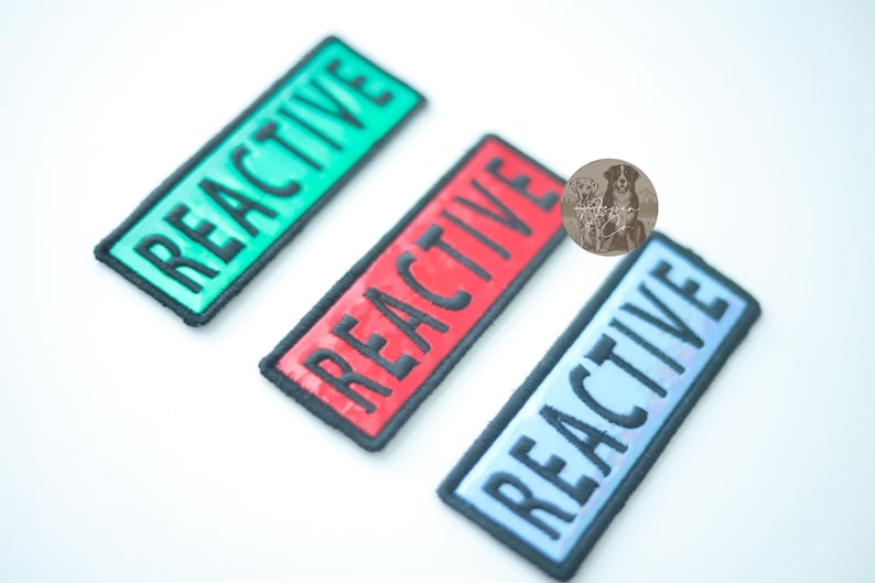 Three holographic REACTIVE dog patches in a 4 x 1.5 inch rectangle shape, shown in red, green, and blue colors. Each patch has hook backing.