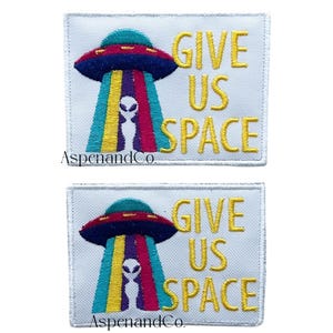 May include: Two embroidered patches with a white background. Each patch features a colorful UFO design with the text "GIVE US SPACE" in yellow. The UFO has a red and blue top, with colorful beams and an alien figure.