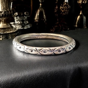 May include: A silver bangle bracelet with a geometric design. The bracelet is made of a thick, round band with a repeating pattern of diamonds and circles.