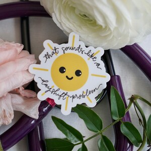 Könnte beinhalten: Ein weißer Aufkleber mit einer gelben Sonnenillustration und dem Text "That sounds like a dayshift problem". Der Aufkleber befindet sich auf einem lila Stethoskop mit Blumen und grünen Blättern.