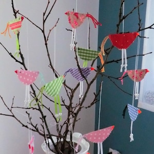 Op de afbeelding: Een witte keramische kan met een bloemenmotief bevat een verzameling kleurrijke stoffen vogels die aan kale takken hangen. De vogels hangen met touw aan de takken en hebben verschillende patronen en kleuren, waaronder rood, roze, groen, blauw en geel.