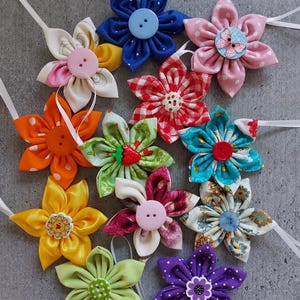 Könnte beinhalten: Eine Sammlung handgefertigter Stoffblumen in verschiedenen Farben, darunter Blau, Rosa, Orange und Gelb. Jede Blume hat eine Knopfmitte und ein weißes Band zum Aufhängen. Die Blumen sind auf einer grauen Oberfläche angeordnet.