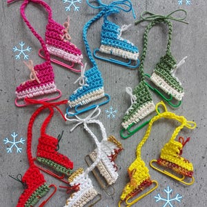 以下が含まれることがあります： ピンク、青、緑、赤、白、黄色など、さまざまな色のミニチュアかぎ針編みのアイススケートのコレクション。各スケートはペーパークリップに取り付けられており、お揃いの糸のループが付いています。スケートは白、金、緑のアクセントで装飾されています。