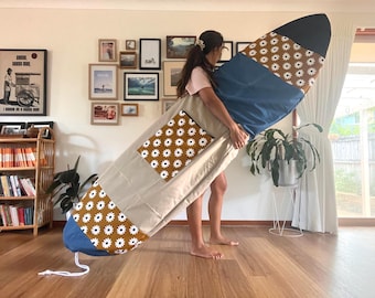 May include: A beige surfboard bag with a blue and brown daisy print. The bag is being held by a person in a room with a bookshelf and other decor.