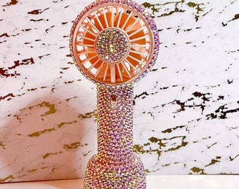 May include: A pink handheld fan with a clear iridescent crystal design. The fan has a round base and a handle with a button on the side.