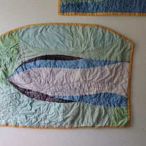 May include: A wall hanging made of patchwork fabric in shades of blue, green, and purple. The fabric is quilted and has a wavy design. The hanging is bordered with a yellow trim.