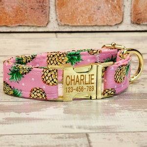 May include: Pink dog collar with a gold pineapple print and a gold buckle. The buckle has the name "CHARLIE" and the phone number "123-456-789" engraved on it.
