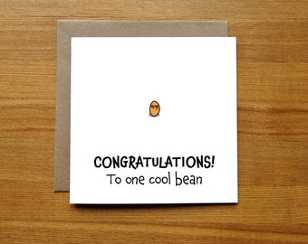 Op de afbeelding: Een witte wenskaart met een bruine envelop. De kaart toont een cartoonboon met een zonnebril en de tekst "CONGRATULATIONS! To one cool bean" in zwarte letters. De kaart ligt op een houten oppervlak.