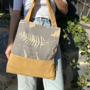 Może przedstawiać: Torba z tkaniny w kolorze szarym i żółtym. Torba ma wzór liścia paproci i jednolite żółte dno. Torba jest trzymana przez osobę ubraną w białą koszulę i niebieskie dżinsy.