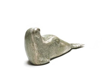May include: A carved stone sculpture of a seal in a reclining position. The sculpture is primarily gray with lighter, natural veining. The seal's body is smooth, with a defined head and tail flipper. The sculpture is set against a white background.
