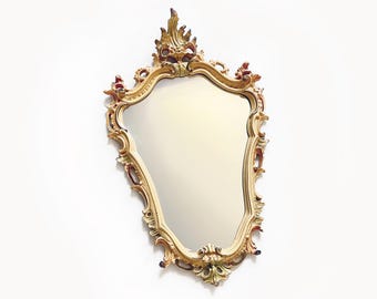 May include: A gold-toned ornate mirror with a scalloped, ornate frame. The mirror has a slightly distressed finish and is shaped like a shield.