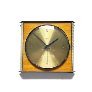 Könnte beinhalten: Eine rechteckige Uhr mit goldfarbenem Zifferblatt und schwarzen Zeigern. Das Zifferblatt ist von einem hellbraunen Holzrahmen und einer dekorativen silbernen Bordüre umgeben. Die Uhr zeigt "Tissot Electronic".