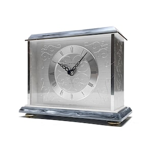Puede incluir: Reloj de cuarzo plateado con forma rectangular. La esfera del reloj presenta números romanos y manecillas negras. El reloj tiene patrones florales decorativos y está sobre una base con patas doradas.