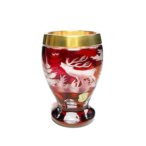 Puede incluir: Una copa de cristal roja con borde dorado. La copa presenta un diseño blanco grabado de un ciervo en una escena de bosque. La base de la copa también es roja y el cristal tiene un acabado brillante.
