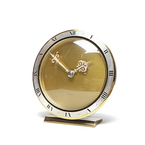 Può includere: Un orologio rotondo color oro con numeri romani e lancette ornate. Il quadrante è di colore oro chiaro e la lunetta è argentata. L'orologio ha una base rettangolare e la parola "DELUXE" è visibile sul quadrante.