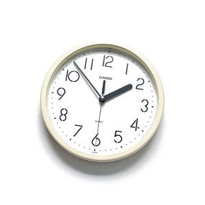 May include: A round, cream-coloured Casio quartz wall clock with a white face. Black numerals and hands mark the time. The clock is approximately 23 cm in diameter.