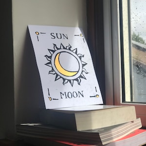 May include: A white card featuring a sun and moon design, with the words "SUN" and "MOON" printed on it, is propped up on a stack of books. The sun has black rays and a yellow crescent moon. The books are on a wooden surface.