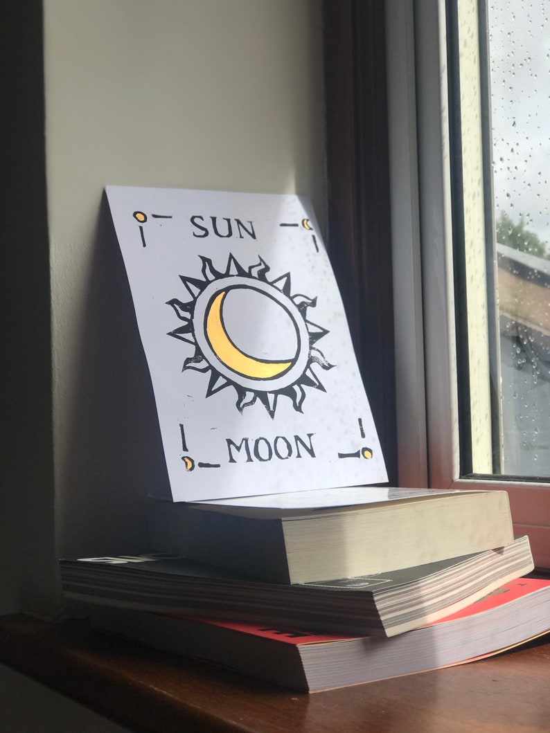 May include: A white card featuring a sun and moon design, with the words "SUN" and "MOON" printed on it, is propped up on a stack of books. The sun has black rays and a yellow crescent moon. The books are on a wooden surface.