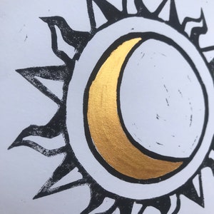 May include: A black and white sun and moon design with a gold crescent moon. The sun has pointed rays, and the moon is centered within the sun's circle. The design is on a white background.
