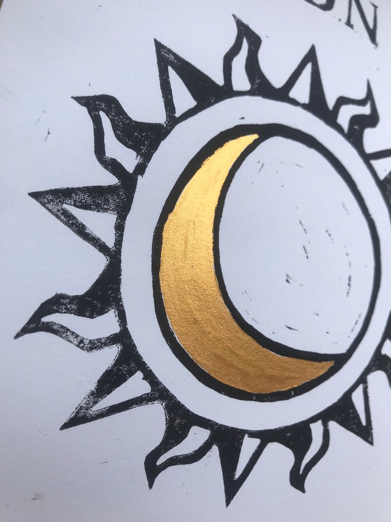 May include: A black and white sun and moon design with a gold crescent moon. The sun has pointed rays, and the moon is centered within the sun's circle. The design is on a white background.