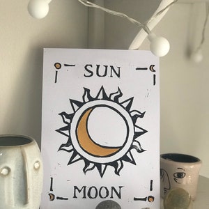 May include: A white print featuring a sun and moon design in black and gold, with the words "SUN MOON". The print is displayed on a white shelf with decorative items, including small ceramic pots and stones. A string of white lights is above the print.