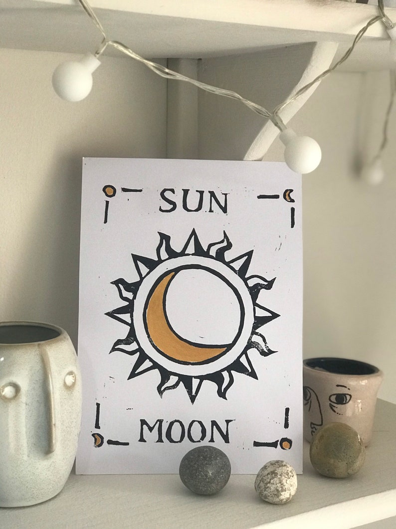 May include: A white print featuring a sun and moon design in black and gold, with the words "SUN MOON". The print is displayed on a white shelf with decorative items, including small ceramic pots and stones. A string of white lights is above the print.