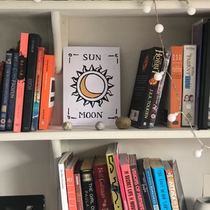 May include: A white bookshelf filled with books, DVDs, and decorative items. A sign with a sun and moon design is centered among the books. String lights with white orbs are draped across the top shelf, adding a soft glow to the scene.