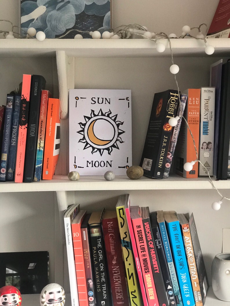 May include: A white bookshelf filled with books, DVDs, and decorative items. A sign with a sun and moon design is centered among the books. String lights with white orbs are draped across the top shelf, adding a soft glow to the scene.